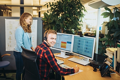 LBS Auszubildender Fachinformatiker sitzt vor zwei Bildschirmen und lächelt in die Kamera. Eine Kollegin guckt dabei auf die Inhalte der Bildschirme