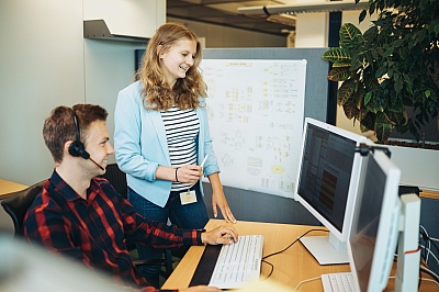 Fachinformatikern für Anwendungsentwicklung erläutert stehend einem sitzenden Fachinformatiker eine Idee auf dem Bildschirm.