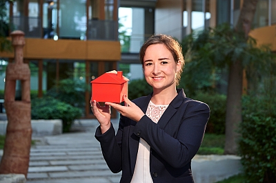 LBS-Kollegin-lächelt-mit-rotem-Haus-in-der-Hand-in-die-Kamera LBS Kollegin lächelt in die Kamera, während sie ein rotes Haus in der Hand hält