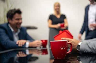 LBS-Beraterinnen-und-Berater-stehen-mit-LBS-Kaffeetassen-um-einen-Tisch-herum LBS-Beraterinnen und Berater stehen mit LBS-Kaffeetassen um einen Tisch herum.